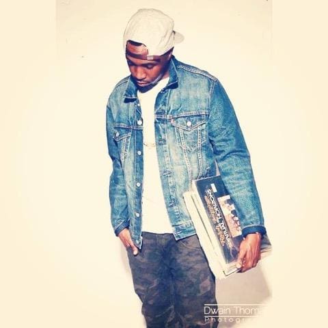 Rashad Thomas wearing a white hat styled backwards, a white shirt, jean jacket and black jeans, holding a collection of vinyl records peering downwards thoughtfully against a yellow background.