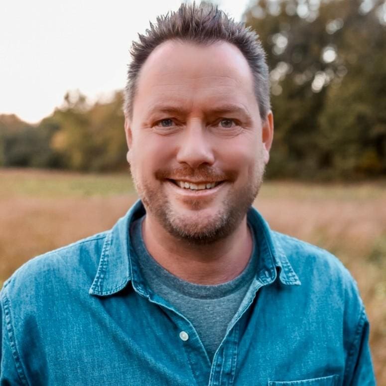 Peter in a jean button down, smiling at the camera, in a field with trees in the background.